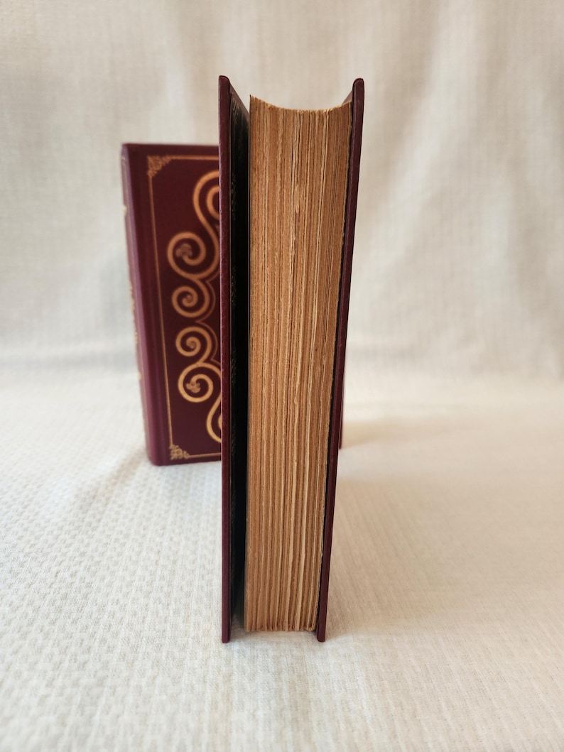 Peut inclure: Deux livres &agrave; couverture rigide bordeaux profond avec des motifs de volutes dor&eacute;s sur les tranches. Les livres sont plac&eacute;s verticalement, r&eacute;v&eacute;lant les bords textur&eacute;s, vieillis et brun clair des pages en papier entre les couvertures sombres.