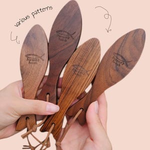 Könnte beinhalten: Eine Sammlung von Massage-Werkzeugen aus Holz mit unterschiedlichen Maserungen. Jedes Werkzeug hat eine ovale Form und ist mit dem Schriftzug "Blessing Brush" und einem Fischsymbol versehen. Sie sind mit einer Lederschnur verbunden. Der Text "various patterns" ist sichtbar.