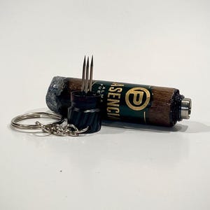 May include: A brown and green cigar-shaped object with the word "ASENCIA" printed on it, along with a "P" logo. A black object with three metal prongs is next to it, along with a silver keychain.
