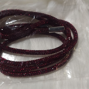 May include: A coiled, burgundy-colored necklace or bracelet, densely covered in sparkling crystals. The jewelry is packaged in a clear plastic bag, with a silver-toned clasp visible. The overall aesthetic suggests a glamorous accessory.