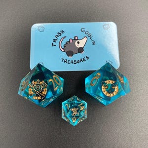May include: A set of three polyhedral dice and a blue box with the text "Trash Goblin Treasures". The dice are teal with gold accents and symbols. The box features a cartoon opossum.