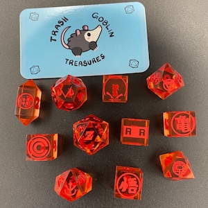 May include: A collection of orange-red dice with red symbols and a blue card with a cartoon possum and the words "Trash Goblin Treasures". The dice are various shapes, including D20s and rectangular blocks.