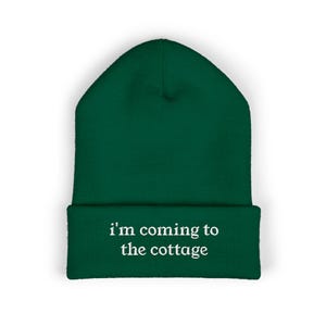 Peut inclure: Un bonnet vert foncé avec le texte blanc "i'm coming to the cottage". Le bonnet est en maille douce et possède un bord replié. Un accessoire décontracté.