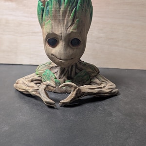 Peut inclure: Sculpture détaillée de Groot des Gardiens de la Galaxie, avec une texture boisée. La figurine a un visage souriant et ses bras forment un cœur. La tête et certaines zones ont des accents verts, ajoutant de la couleur à la base brune.