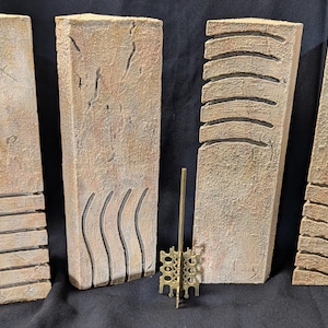 Op de afbeelding: Vier beige rechthoekige stenen pilaren met gegraveerde ontwerpen, een met een gouden decoratieve metalen standaard ervoor.