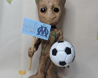 May include: A green and brown Groot figurine holding a soccer ball and a sign that says "Allez L'OM!" in French. The sign also has the logo of the Olympique de Marseille soccer club.