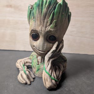 Puede incluir: Un macetero de cerámica verde y marrón con forma de personaje de la película Guardianes de la Galaxia. El personaje está sentado con la barbilla apoyada en la mano.