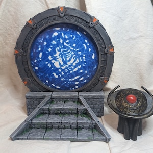 Peut inclure: Une réplique de la Porte des étoiles en gris et orange avec un effet de portail bleu tourbillonnant. La Porte des étoiles est sur un socle en pierre avec des marches menant à elle. Une plus petite réplique de la Porte des étoiles en noir et or avec un centre rouge est au premier plan.