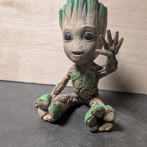 Può includere: Una statuetta dettagliata di Baby Groot da Guardiani della Galassia. La figura è principalmente marrone con accenti verdi sulla testa, sulle spalle e sui piedi. Groot è seduto, con una mano alzata in un saluto.