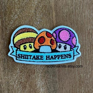 May include: Embroidered patch with three colourful mushrooms and the text "SHIITAKE HAPPENS" on a light blue banner. The mushrooms are brown, orange, and purple with various patterns. The patch is on a wooden surface.