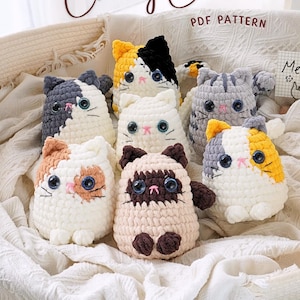 Könnte beinhalten: Eine Sammlung von sieben gehäkelten Katzenplüschtieren in verschiedenen Farben und Mustern, darunter dreifarbig, schwarz-weiß, grau-weiß und braun-weiß. Die Plüschtiere sitzen in einem weißen Weidenkorb mit einer weißen Decke.