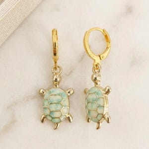 Könnte beinhalten: Ein Paar goldfarbene Creolen, jeweils mit einem kleinen Schildkrötenanhänger. Die Schildkrötenanhänger haben ein hellblaues und weißes Schalendesign. Die Ohrringe werden auf einer weißen Marmoroberfläche präsentiert.