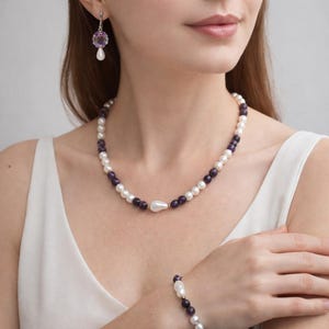 Puede incluir: Un conjunto de collar, pulsera y pendientes con perlas blancas y cuentas moradas oscuras. El collar tiene un colgante de perla grande en forma de lágrima. Los pendientes tienen una gema morada y una gota de perla blanca.