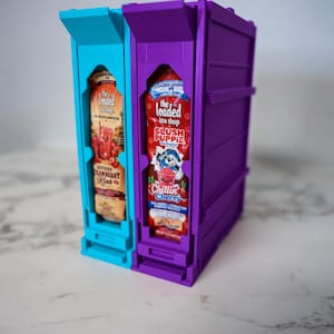 Puede incluir: Un estante de almacenamiento de plástico azul brillante y morado que contiene dos paquetes de mezcla para bebidas. El paquete izquierdo es naranja con la etiqueta The Loaded Tea Shop Southern Cranberry Kiss. El paquete derecho es rojo con la etiqueta The Loaded Tea Shop Slush Puppie Chillin Cherry.