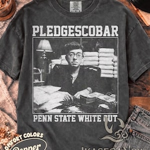 Könnte beinhalten: Ein anthrazitfarbenes Kurzarm-T-Shirt mit einem schwarz-weißen Grafikdruck einer Person, die an einem Schreibtisch mit Papier- und Geldstapeln sitzt. Der Text PLEDGESCOBAR steht über dem Bild und PENN STATE WHITE OUT steht unter dem Bild.