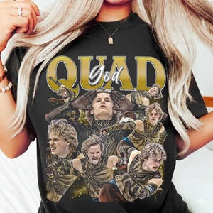 Puede incluir: Camiseta negra con un gráfico dorado que dice "QUAD God" y presenta múltiples imágenes de una persona con un disfraz. Las imágenes están en un estilo de collage, con la persona en varias poses.