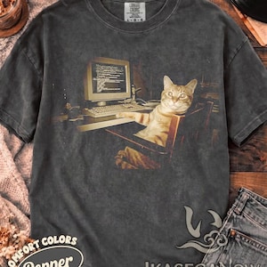 May include: A charcoal gray short-sleeve t-shirt featuring a graphic print of a tabby cat sitting in a chair in front of a vintage computer monitor displaying lines of code. The shirt has a crew neckline and a faded, washed-out fabric appearance.