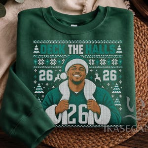 Op de afbeelding: Een donkergroene sweater met ronde hals en een grafische print van een persoon met een kerstmuts en een shirt met het nummer 26. Het ontwerp bevat de tekst DECK THE HALLS, sneeuwvlokken, dennenbomen en rendieren in wit en lichtgroen.