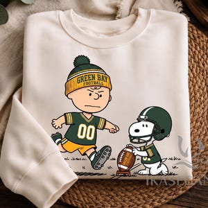 Könnte beinhalten: Ein cremefarbenes Sweatshirt mit Rundhalsausschnitt und einer Grafik eines Zeichentrickcharakters, der eine gelb-grüne Green Bay Football-Mütze und ein Trikot trägt und auf einen Football zuläuft, der von einem Hund mit grünem Helm gehalten wird.