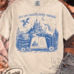 Puede incluir: Una camiseta de manga corta color marfil con un estampado gráfico azul. El diseño incluye un castillo, un dragón, un unicornio, libros apilados, una taza de té y flores. El texto "it's too nonfictiony outside" aparece sobre la ilustración.