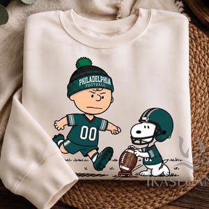 Könnte beinhalten: Ein cremefarbenes Sweatshirt mit Rundhalsausschnitt und einer Grafik eines Zeichentrickcharakters, der eine grüne Philadelphia Football-Mütze und ein Trikot trägt und auf einen Football zuläuft, der von einem Hund mit Helm gehalten wird.