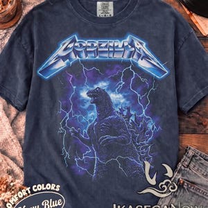 May include: A navy blue short-sleeve t-shirt featuring a graphic print of Godzilla standing amidst purple and blue lightning bolts. The word Godzilla is printed in a metallic blue font above the creature. The shirt has a crew neckline and a tag.