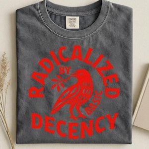 Peut inclure: T-shirt gris foncé avec un motif rouge. Le motif représente un oiseau tenant une branche dans un cercle, avec les mots "RADICALIZED BY BASIC DECENCY" en rouge. Le t-shirt a un col rond et une étiquette Comfort Colors.