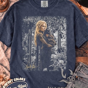 May include: A navy blue short-sleeve t-shirt featuring a graphic print of a person with long blonde hair holding a cat in a garden setting. The print includes a signature in the bottom left corner. The shirt has a crew neck and a vintage wash finish.