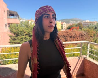 May include: A woman wearing a burgundy and gold patterned headscarf with long tassels. The headscarf is draped over the head and shoulders. She is wearing a black top. The background includes buildings and greenery.