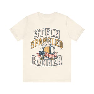 Pode incluir: Uma t-shirt de manga curta na cor creme com um gráfico de uma caneca de cerveja com espuma, uma fita com a bandeira americana e lúpulo. O texto STEIN SPANGLED BANNER está escrito em letras de bloco em negrito dispostas em arco.