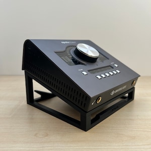 Puede incluir: Una interfaz de audio negra con una perilla grande y una pantalla, sentada en un soporte negro. La interfaz está etiquetada como "Apollo Twin" y "Universal Audio".