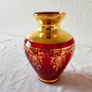 May include: A small ruby red glass vase featuring a wide, polished gold-tone neck and a matching gold-tone base. The body of the vase is decorated with an intricate, repeating gold filigree pattern that encircles the midsection of the glass.