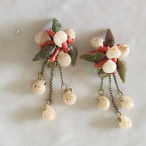 May include: A pair of clip-on earrings featuring clusters of small white and green seashells accented with bright orange coral-like pieces. Each earring has three delicate gold-tone metal chains dangling with small white spiral seashells at the ends.