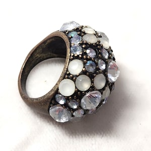 Puede incluir: Un anillo de banda ancha con acabado en bronce antiguo, que presenta una superficie abovedada incrustada con un grupo de cristales facetados transparentes y piedras cabujón blancas opacas y lisas de varios tamaños en un patrón denso.