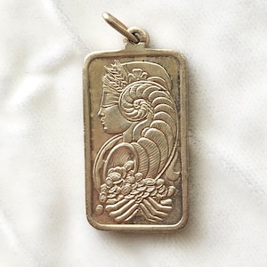 Peut inclure: Pendentif rectangulaire en argent représentant le profil d'une femme avec un bandeau sur les yeux et une coiffure élaborée. Le pendentif comporte une boucle pour une chaîne et un motif floral.