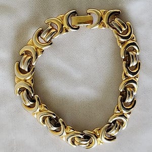 May include: Gold-tone bracelet with a repeating link design. The bracelet features a series of interlocking, crescent-shaped links in a gold color, with silver-colored accents. The clasp is a simple, rectangular design. The bracelet is displayed on a white surface.