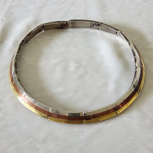 May include: A rigid collar necklace constructed from interlocking curved segments. The design features three distinct horizontal bands of metal in silver, copper, and gold tones, secured with a hinged clasp mechanism at the back of the neck.