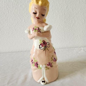 May include: A ceramic figurine of a person with blonde hair, wearing a peach-colored dress with white accents and a floral pattern of purple flowers and green leaves. The figure has closed eyes, painted facial features, and holds a small bouquet.