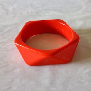 May include: A bright orange bangle bracelet with a distinct geometric, hexagonal shape. The accessory features a smooth, solid-colored surface with sharp, angled edges that form a six-sided perimeter around the central circular opening.