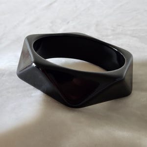 May include: A solid black bangle bracelet with a smooth, glossy finish. The bracelet features a distinct geometric design with multiple faceted, angular sides that create a sharp, multi-sided shape around the circular opening for the wrist.