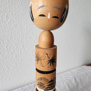 Può includere: Bambola Kokeshi giapponese in legno. La testa rotonda ha capelli neri dipinti, occhi chiusi e una piccola bocca rossa. Il corpo cilindrico è decorato con scene dipinte in bianco e nero della natura e dell'architettura.