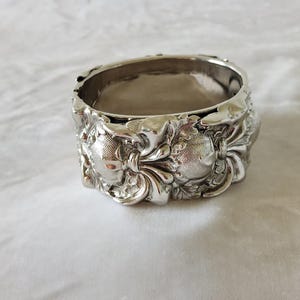 Puede incluir: Un servilletero de metal plateado con un patrón en relieve muy detallado de motivos redondeados y texturizados similares a flores o frutas, intercalados con elementos lisos y curvos en forma de hoja que rodean todo el anillo circular.
