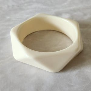 May include: A solid off-white bangle bracelet featuring a distinct geometric hexagonal shape with smooth, angled edges and a polished finish. The rigid circular band has a wide profile and a uniform cream-colored surface throughout the entire piece.