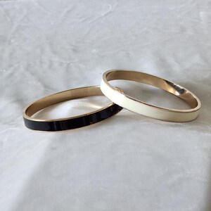 May include: Two circular bangle bracelets with a smooth, polished finish. One bracelet features a glossy black enamel inlay, while the other features a glossy cream-colored enamel inlay. Both bangles have a metallic gold-toned interior and edge.