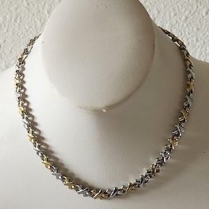 May include: A two-tone necklace featuring a repeating pattern of alternating silver-colored and gold-colored X-shaped links. The chain is constructed from interconnected metal segments that create a continuous, textured design around the entire length.