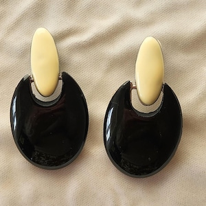 May include: A pair of statement earrings. Each earring features a glossy black, crescent-shaped base with a cream-colored oval accent at the top. The earrings have a vintage aesthetic and are set against a neutral background.