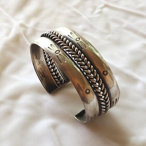 May include: A silver cuff bracelet featuring a central braided rope texture flanked by two smooth bands. The outer bands are decorated with stamped circular and elongated oval patterns. The metal has a polished finish with darkened recessed areas.