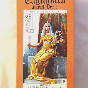 Puede incluir: Una caja naranja de Cagliostro Tarot Deck con el texto "Cagliostro Tarot Deck" en la parte superior. La ilustración muestra una figura con una túnica amarilla sosteniendo una espada y una balanza, con el texto "JUSTICE UNIVERSAL BALANCE - DIVISION JUSTICE" debajo.