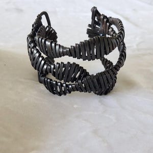 Könnte beinhalten: Ein dunkelbraunes Armband in Manschettenform, das aus mehreren Drahtsträngen besteht, die in einem durchgehenden, sich wiederholenden Spulenmuster gewickelt sind. Das Design weist eine geschichtete, offene Struktur mit metallischem Glanz auf.