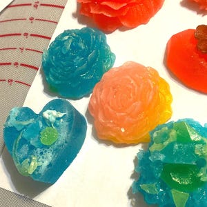 May include: An assortment of colorful, handcrafted candies in various shapes. The image features candies shaped like roses, hearts, and abstract forms, in shades of orange, blue, and green. The candies are arranged on a white surface.